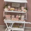 Charrette blanche Candy Bar décorée avec bocaux de bonbons, cupcakes, macarons, cake pops, fleurs blanches et roses, disponible à la location pour événements.