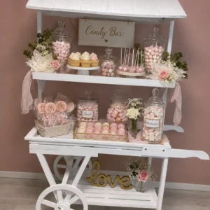 Charrette blanche Candy Bar décorée avec bocaux de bonbons, cupcakes, macarons, cake pops, fleurs blanches et roses, disponible à la location pour événements.