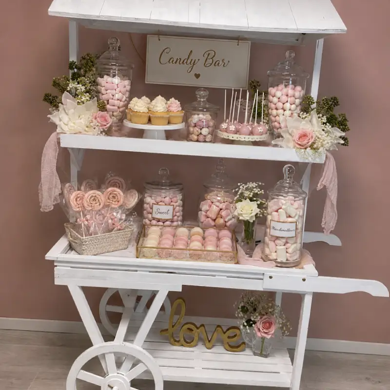 Charrette blanche Candy Bar décorée avec bocaux de bonbons, cupcakes, macarons, cake pops, fleurs blanches et roses, disponible à la location pour événements.