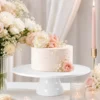 Location présentoir à gâteau blanc Présentoir à gâteau blanc avec gâteau fleuri, fleurs rose poudré et blanches, bougies et macarons dans une décoration événementielle raffinée.