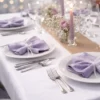 Table de réception élégante avec serviettes de table lilas pliées sur des assiettes blanches, verrerie fine, bougie et composition florale.