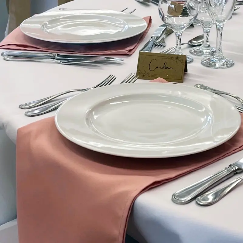 Table de réception avec serviettes de table rose poudré, assiettes blanches, couverts argentés et verres élégants.