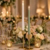 Deux bougies flambeaux LED haut de gamme, blanche et vert sauge, allumées dans des bougeoirs dorés sur une table de mariage élégante avec fleurs et verrerie.