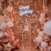 Décor d’anniversaire avec mur de sequins rose gold, néon Happy Birthday, ballons rose gold, blanc et nude, et support transparent avec gâteau.