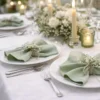 Table de réception élégante avec serviettes de table vert sauge pliées sur des assiettes blanches, décor floral et bougies.