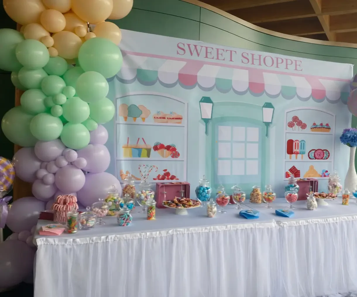 Candy bar décoré avec une arche de ballons pastel et une table de douceurs devant un fond Sweet Shoppe.