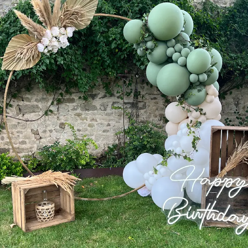 Décoration d’anniversaire bohème avec arche de ballons vert sauge, beige et blanc, cercle doré et éléments naturels en extérieur.