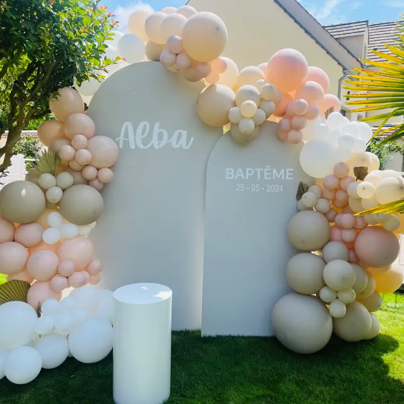 Décoration de baptême avec panneaux personnalisés, ballons organiques blanc, beige et rose poudré dans un jardin.