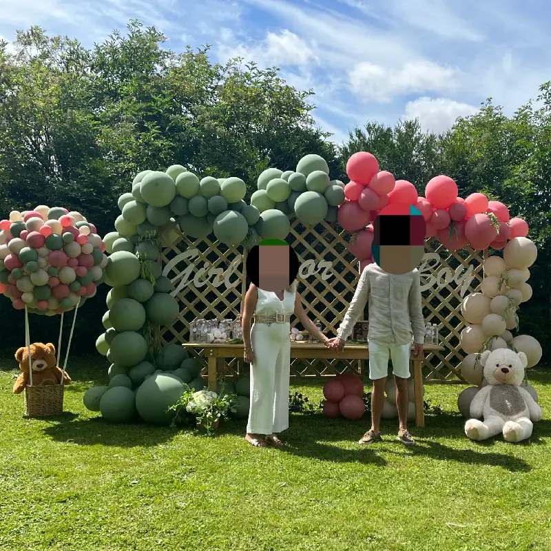 Décoration de gender reveal en extérieur avec arche de ballons vert sauge, rose et beige, table décorée et éléments scénographiques.