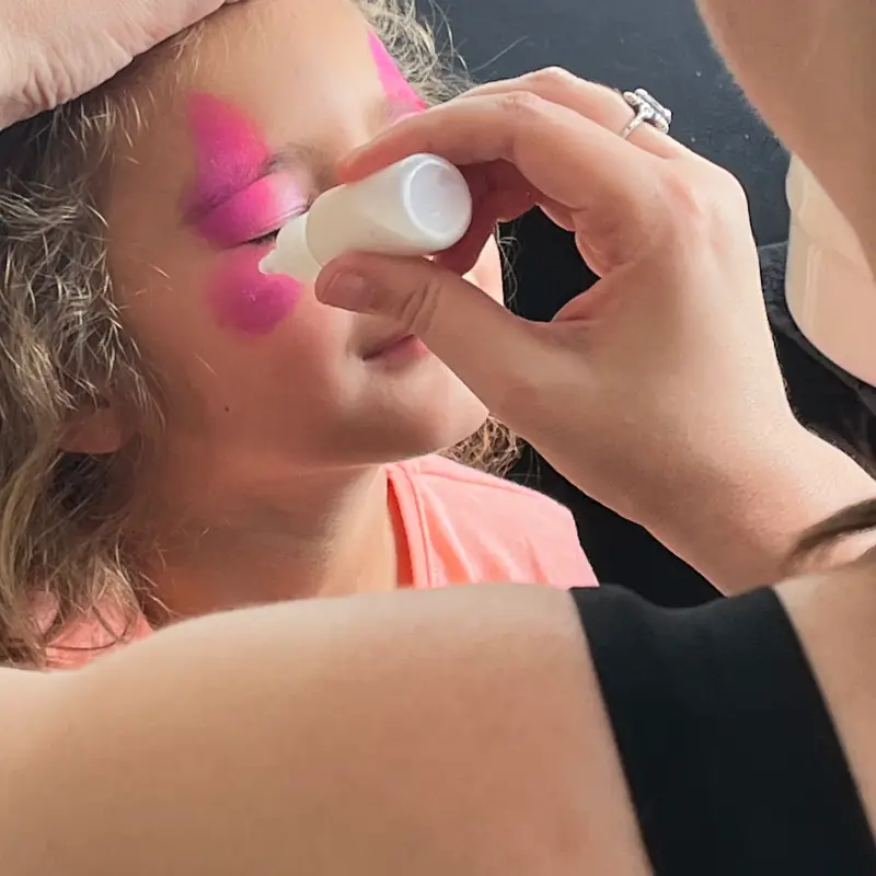 Animatrice réalisant un maquillage rose sur le visage d’une enfant lors d’une kermesse.
